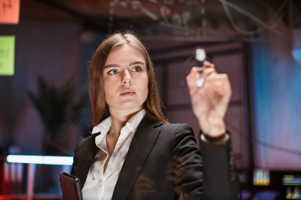 Young focused woman writing on glass office board in modern office